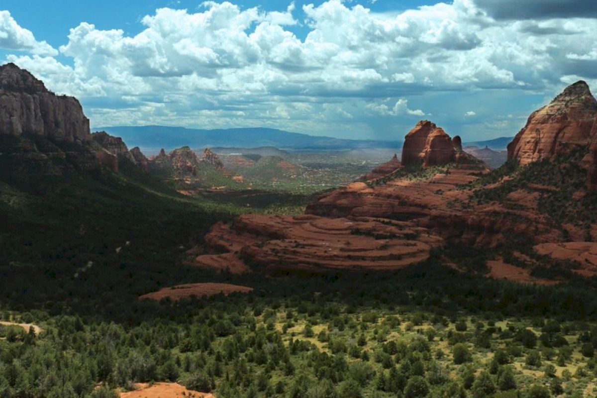 A scenic landscape of red rock formations and green forest, with a vast sky overhead filled with clouds in the background.