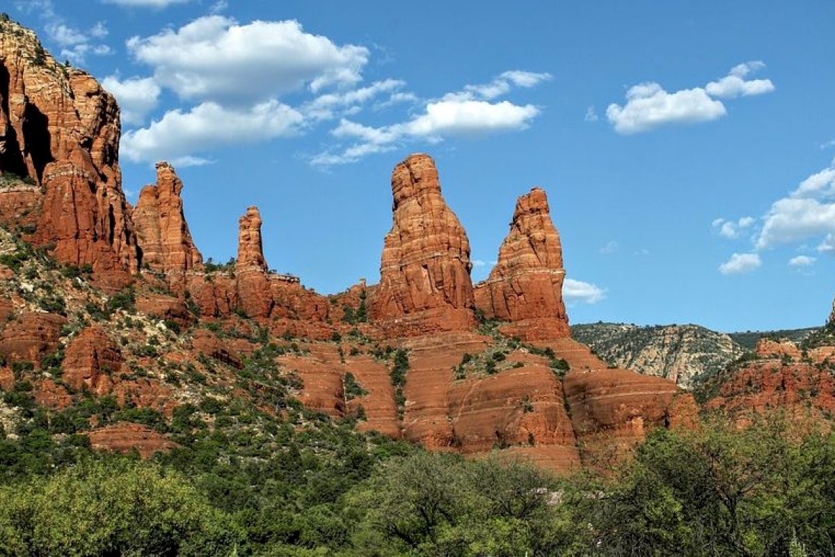 The image shows a landscape with striking red rock formations under a blue sky with scattered clouds, and green vegetation in the foreground.