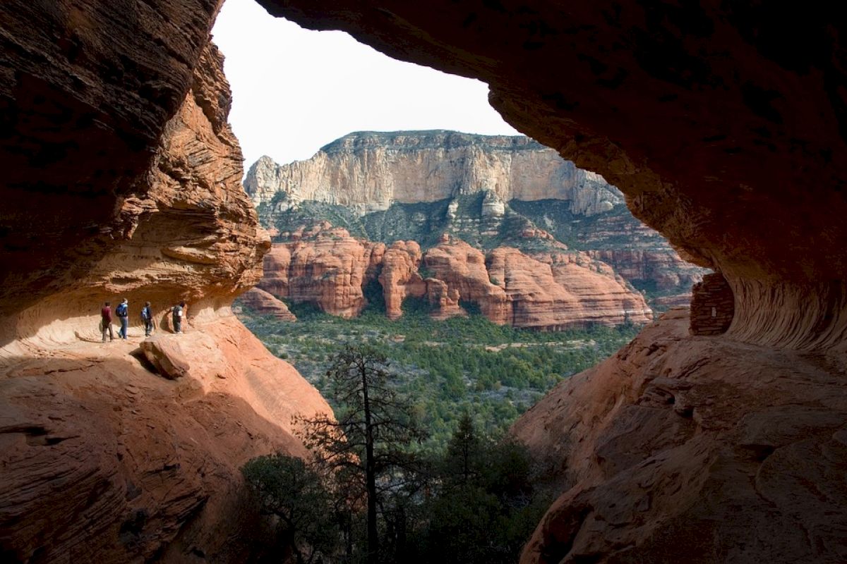 A group of people are hiking through a large rock formation, enjoying a scenic view of red rock cliffs and greenery in the background.