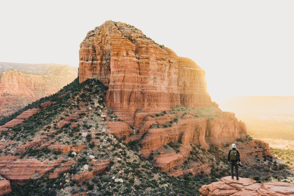 A lone hiker stands on a rocky plateau, overlooking a vast desert landscape with a large, sunlit red rock formation in the background.