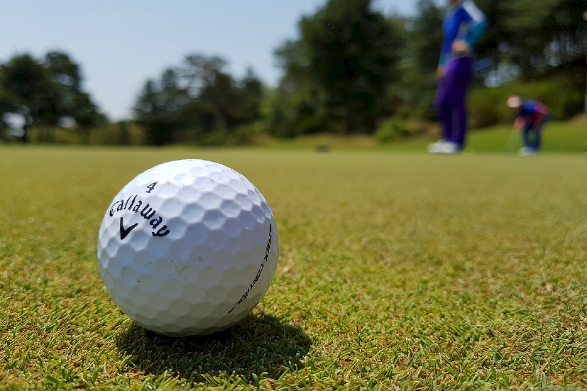 A close-up of a Callaway golf ball on a golf course green, with two golfers in the background preparing to putt, surrounded by trees and greenery.