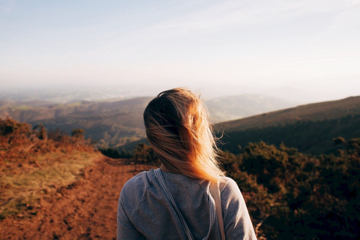 A person with blonde hair stands on a dirt path, overlooking a scenic mountain landscape under a clear sky during the day.