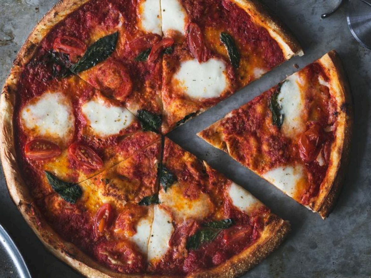 The image shows a pizza with melted cheese, tomato sauce, and basil leaves on a baking tray. A pizza cutter is next to it, with one slice partially removed.