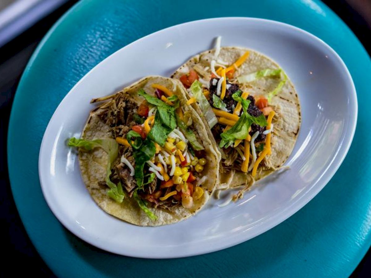 The image shows a white plate with two tacos topped with shredded cheese, lettuce, diced tomatoes, and other ingredients on a teal surface.