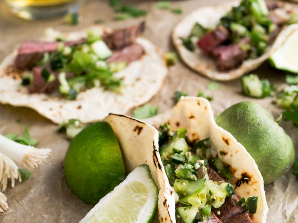 The image shows several tacos with sliced beef, fresh greens, and lime wedges on a wooden surface. There is also a glass of beverage and some lime.