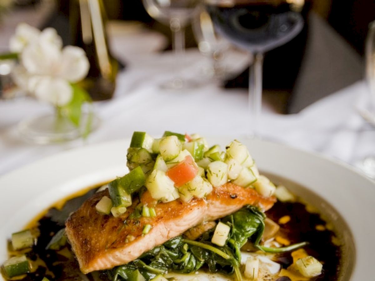 A gourmet dish featuring a seared piece of fish topped with a vegetable medley, plated on a bed of greens, with wine glasses in the background.