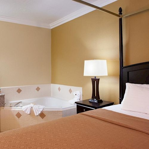 A cozy hotel room with a bed, bedside lamp, bathtub, vanity, and a mirror. The room has a warm color palette with white and brown tones.