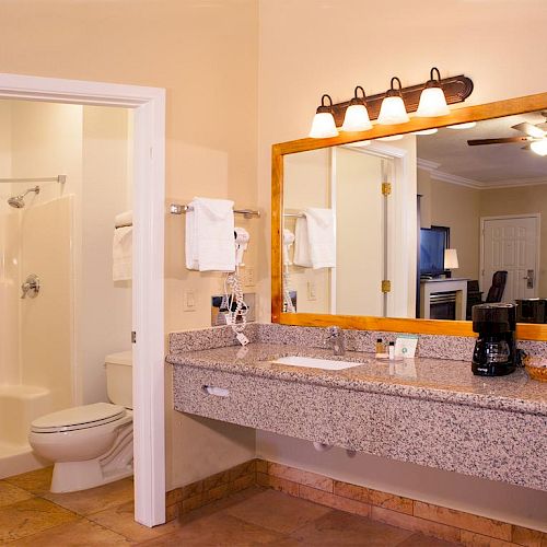 The image shows a bathroom with a large mirror, sink, and vanity, connected to a room with a bed, TV, and ceiling fan.