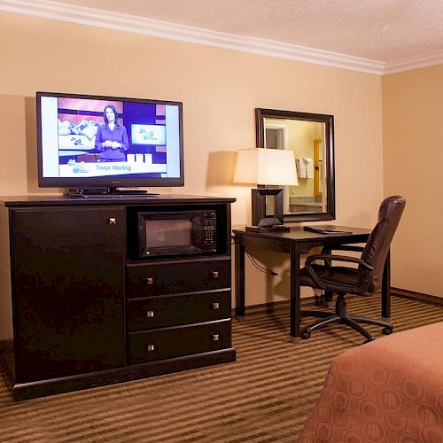 A cozy hotel room with a TV, dresser, microwave, desk, chair, lamp, and a visible bathroom area, all atop a carpeted floor.