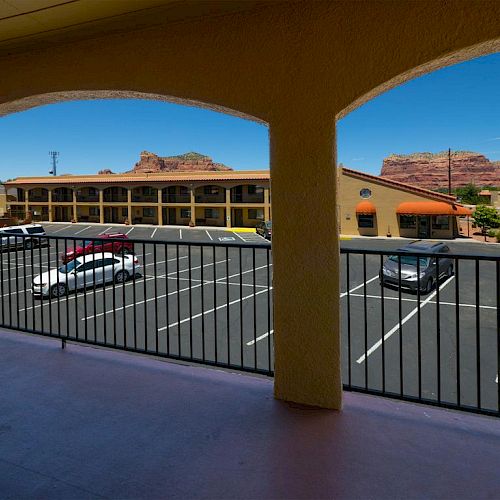 A view from a covered balcony shows a parking lot with several cars and a motel building, against a backdrop of rocky terrain under a clear sky.