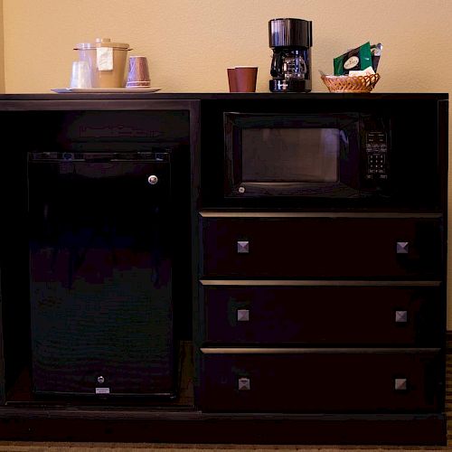 The image shows a cabinet with a mini-fridge, a microwave, three drawers, a coffee maker, coffee cups, and condiments on top.