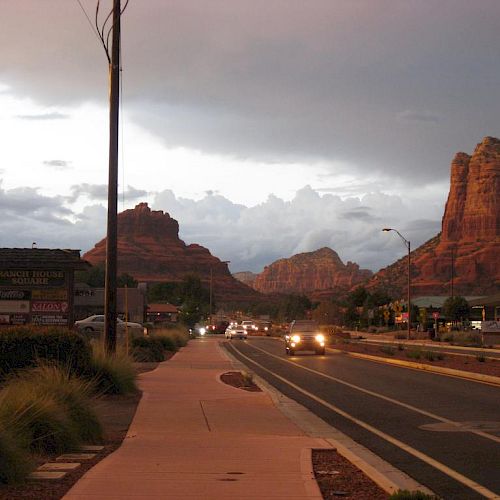 A scenic road in a desert town during sunset, featuring distinctive red rock formations and a few vehicles with headlights on.