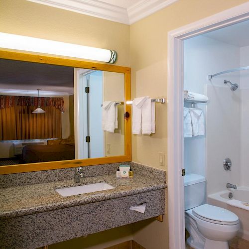 The image shows a hotel bathroom with a granite countertop, mirror, sink, towel rack, toilet, and a shower-tub combo in the adjoining room.