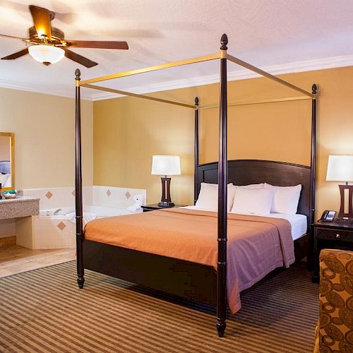 A cozy bedroom features a four-poster bed, a jacuzzi tub, two nightstands with lamps, a ceiling fan, and a vanity area with a mirror and sink.