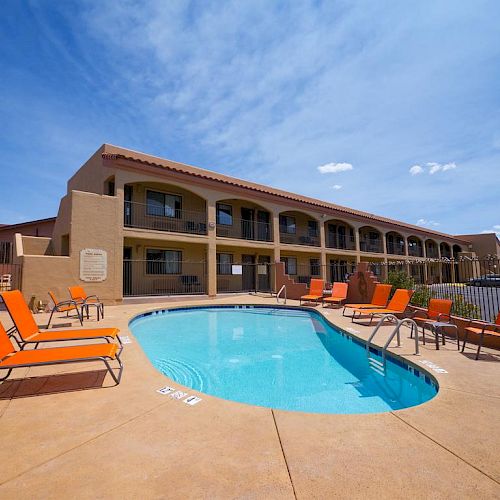 Image of a hotel pool area with an adjacent outdoor seating area. There are sun loungers around the pool and a two-story hotel building in the background.