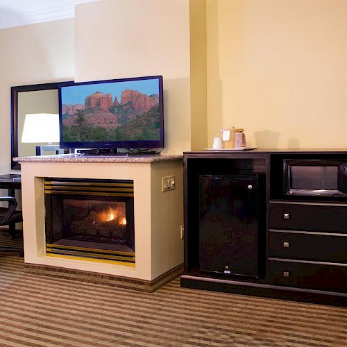 The image shows a cozy room with a television above a fireplace, a black cabinet featuring a mini fridge and microwave, and a work desk with a chair.