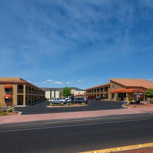 A roadside hotel with a stucco exterior, orange awnings, and a parking lot in front, surrounded by desert landscaping and under a blue sky.