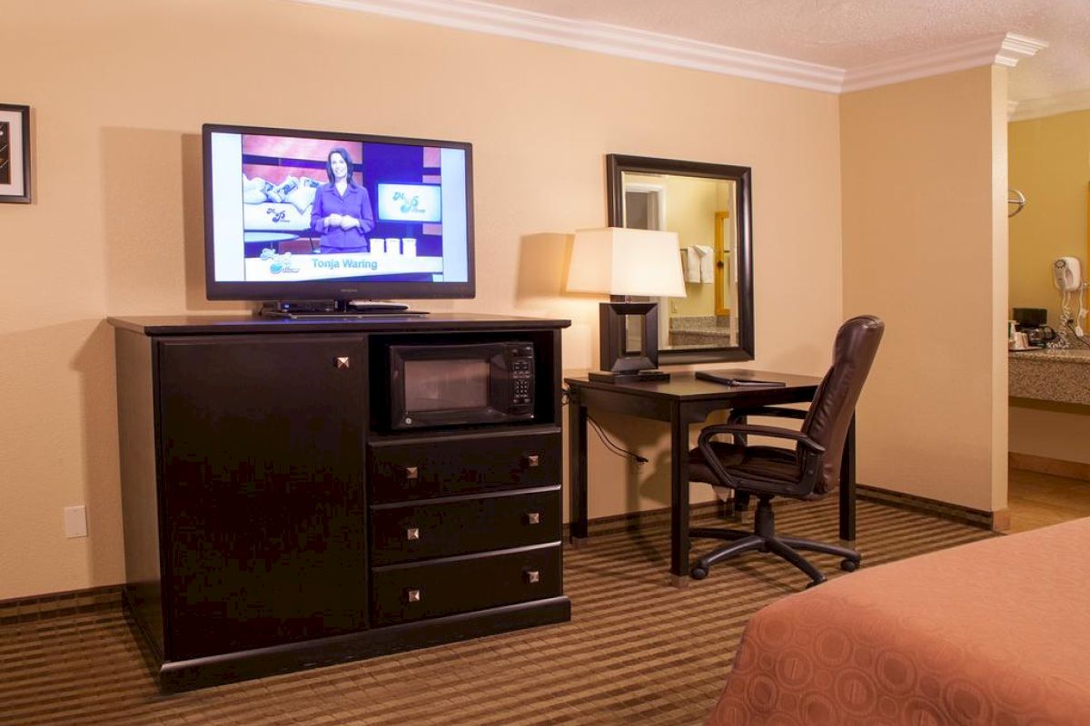 A hotel room features a TV, microwave, dresser, desk with a lamp, mirror, office chair, and part of a bed with a brown bedspread.