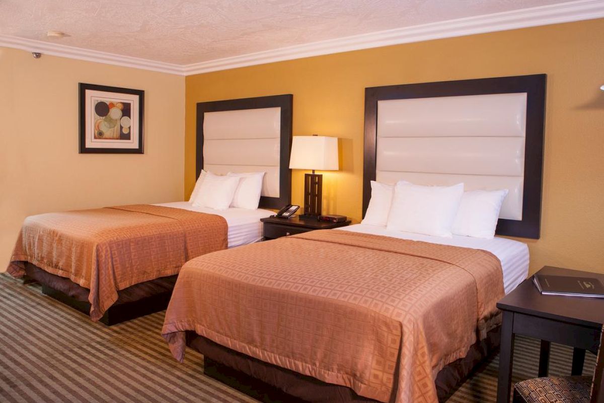 The image shows a hotel room with two neatly made double beds, a nightstand with a lamp, a striped carpet, and a framed picture on the wall.