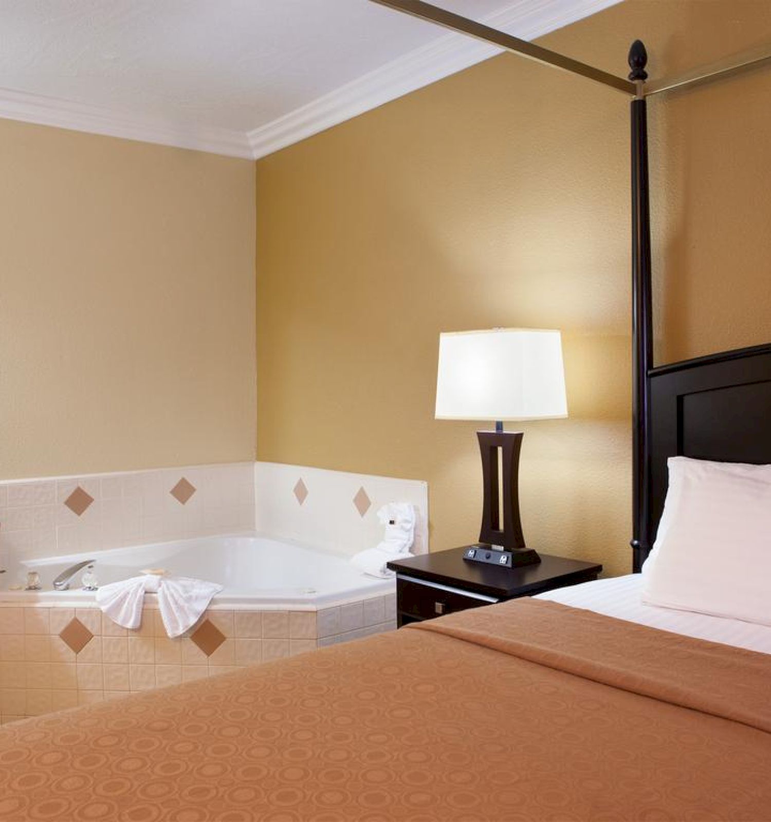 This image shows a hotel room with a large bed, a bedside table with a lamp, and a jacuzzi tub next to a small countertop with a sink and mirror.