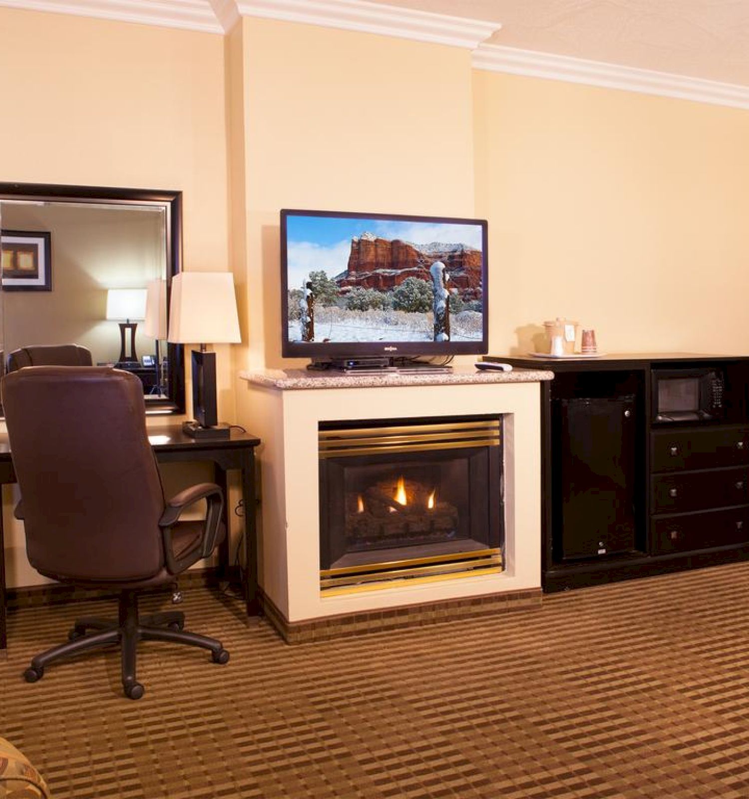 A cozy hotel room features a fireplace, desk with a chair and lamp, a flat-screen TV, a dresser, a microwave, and a wardrobe.