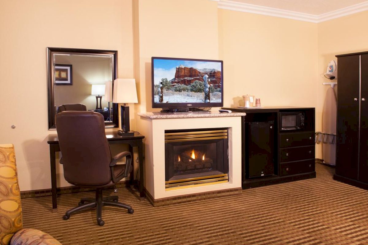 A cozy hotel room with a fireplace, flat-screen TV, desk, chair, lamp, mirror, and a cabinet with a microwave and mini-fridge.