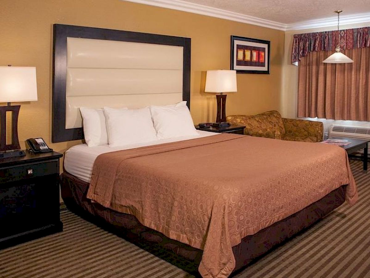 The image displays a hotel room with a large bed, two nightstands with lamps, a sofa, a window with curtains, a framed picture, and striped carpet.