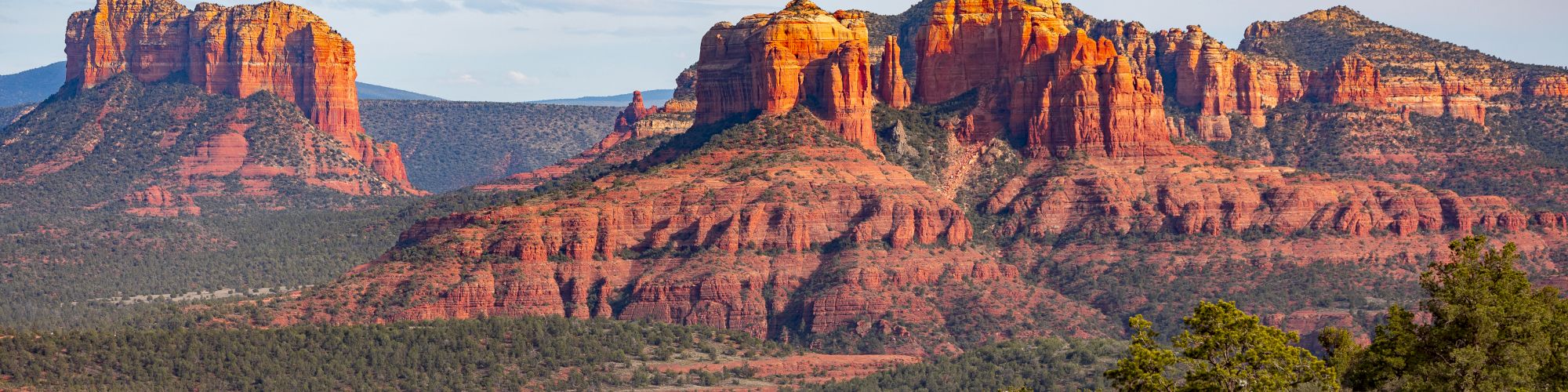 A scenic view of towering red rock formations and mesas amidst a semi-arid landscape, under a partly cloudy sky with green vegetation in the foreground.