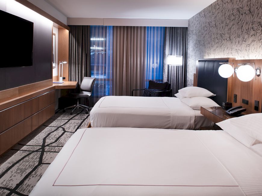 A modern hotel room with two beds, a flat-screen TV, desk, and chair, featuring stylish decor and ambient lighting.