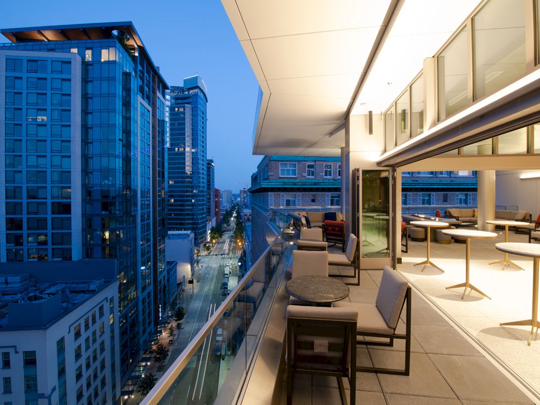A modern rooftop balcony with tables and chairs overlooks a city street with tall buildings at dusk, featuring glass and steel architecture.