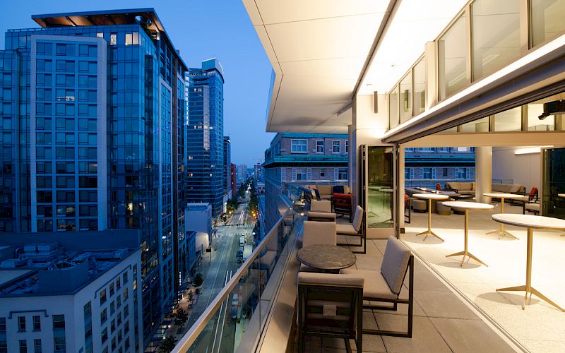 A modern rooftop terrace overlooking a cityscape with tall buildings, featuring outdoor seating and tables, illuminated in the evening light.