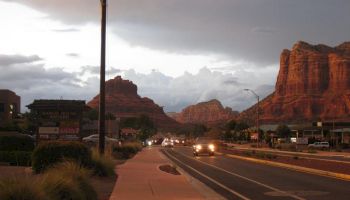 A scenic road with cars, streetlights, and buildings set against a backdrop of striking red rock formations under a cloudy sky.