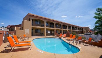 A small outdoor swimming pool with orange lounge chairs, located in front of a two-story building under a clear blue sky, is shown in the image.