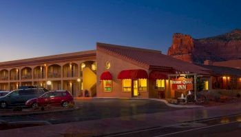 The image shows a well-lit inn with a sign reading "Desert Quail Inn," red awnings, parked cars, and a rocky landscape in the background during dusk.