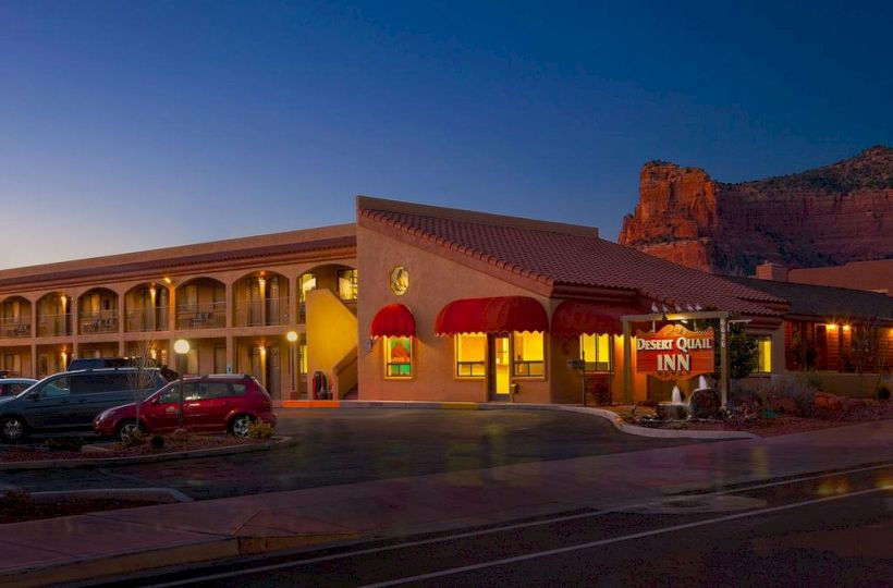 The image depicts a well-lit hotel named "Desert Quail Inn" at dusk, with parked cars in front and a rocky landscape in the background.