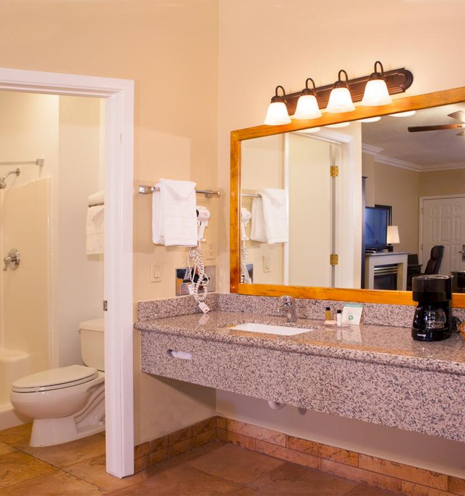 The image shows a bathroom with a large mirror, a countertop with a coffee maker, and an adjoining room with a toilet and shower.