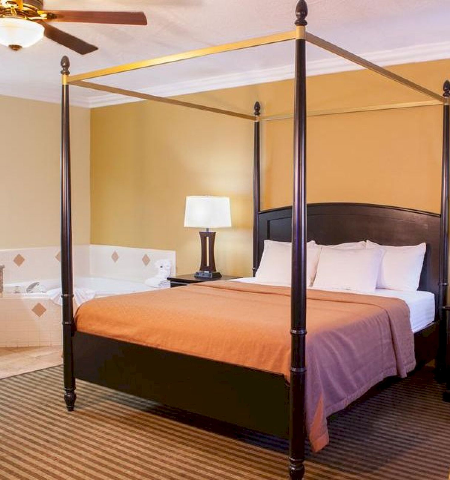 A well-lit hotel room with a four-poster bed, armchair, bedside tables, lamps, a whirlpool tub, a vanity area with a mirror, and a ceiling fan.