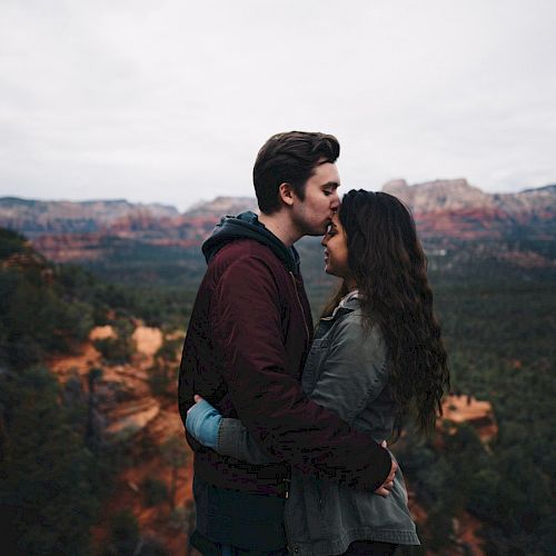 A couple embraces on a cliffside with scenic mountains and forests in the background, the man affectionately kissing the woman's forehead.