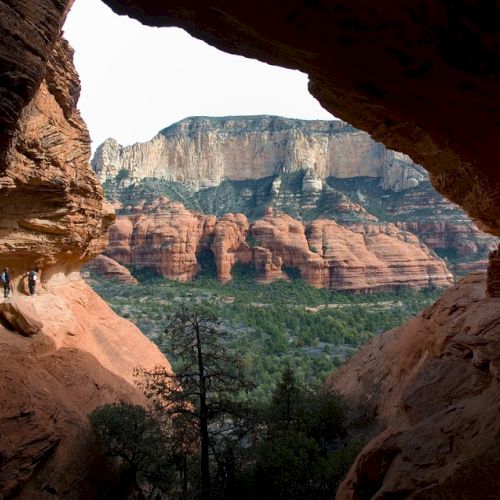 People hiking through a cavernous rock formation, with a view of a stunning red rock landscape and towering cliffs in the background.