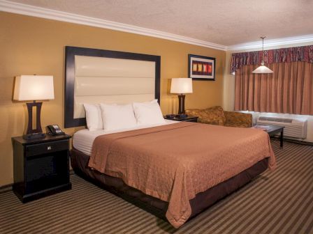 This image shows a hotel room with a large bed, two bedside tables with lamps, a sofa, a painting, and a window with curtains and a hanging light.