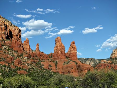 This image shows a scenic view of red rock formations against a bright blue sky with scattered clouds, surrounded by green vegetation.